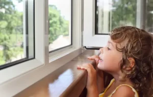 A young child with curly hair eagerly looks out an open window, smiling at the view outside on a sunny day.